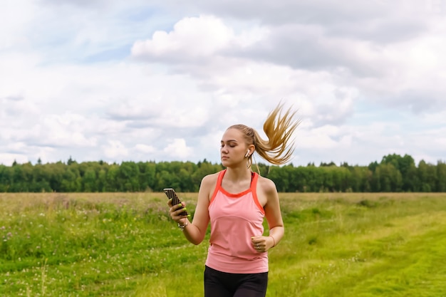 Young woman listening to music or reads messages while jogging along a path through a meadow