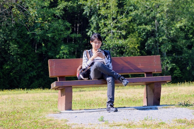 Photo young woman listening music through smart phone while sitting on bench against trees at park