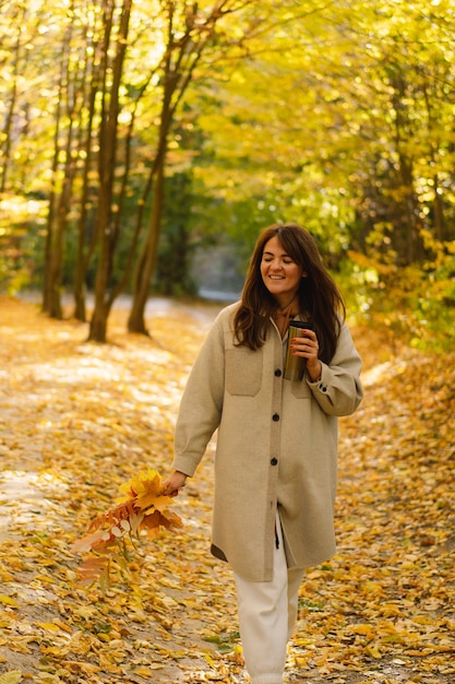 A young woman in a long shirt with a thermo mug in her hands walks through the autumn forest