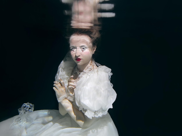 young woman in long white luxury dress underwater on the black background. Motion, art, winter