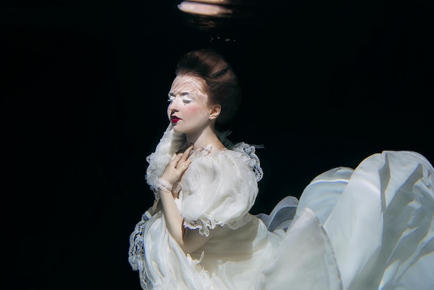 young woman in long white luxury dress underwater on the black background. Motion, art, winter