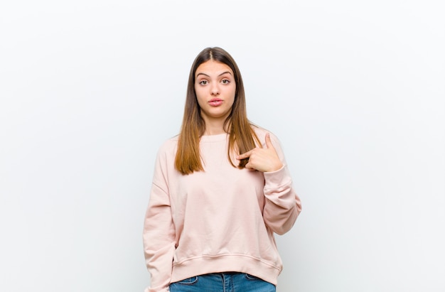 Young woman looking happy, proud and surprised, cheerfully pointing to self, feeling confident and lofty on white