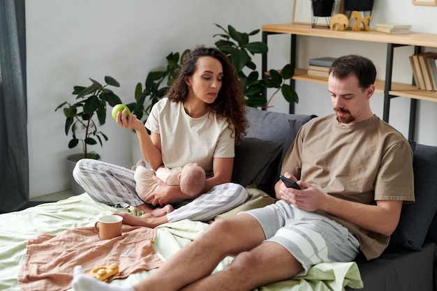 Young woman looking at screen of smartphone of her husband sitting next to her