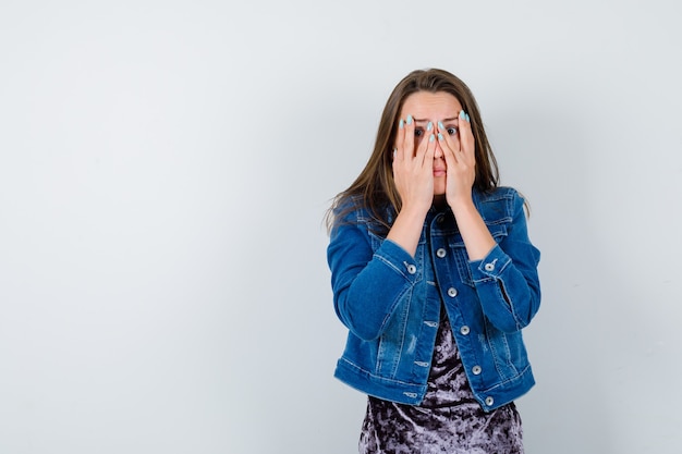 Young woman looking through fingers in denim jacket and looking frightened , front view.