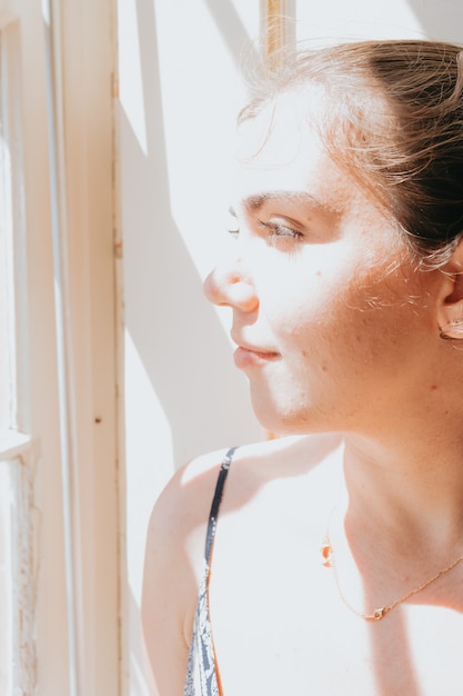 Young woman looking through a window during a super sunny day, copy space, reflexion and thinking concept, sad and anxiety, self care