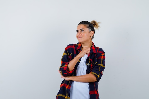 Young woman looking up, with hand near face in checkered shirt and looking dissatisfied , front view.