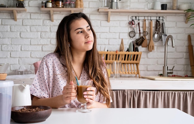 Young woman in lovely pajamas drinking coffee at home kitchen