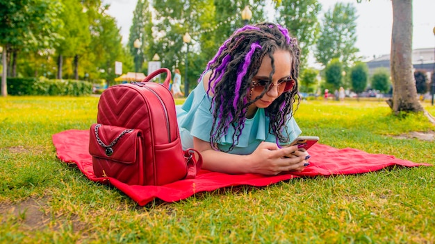 Young woman lying on a blanket blowing uses a smartphone in the park Happy young brunette female lying on the grass and looking through the phone sunny day