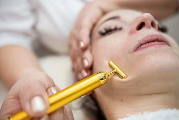 Young woman lying on cosmetologist's table and applying procedure with vibration massager on face Skincare lifting at spa