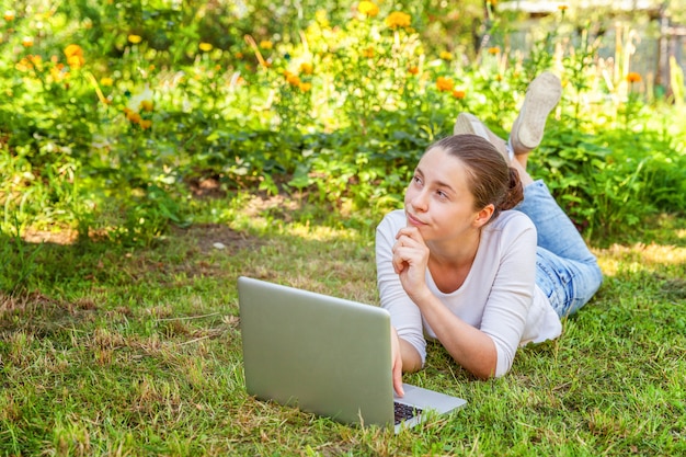 Young woman lying on green grass lawn in city park working on laptop pc computer.