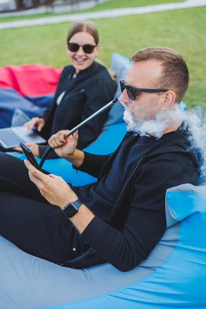 A young woman and a man are sitting in a park on a bean bag and smoking hookah Happy couple relaxing outdoors with hookah