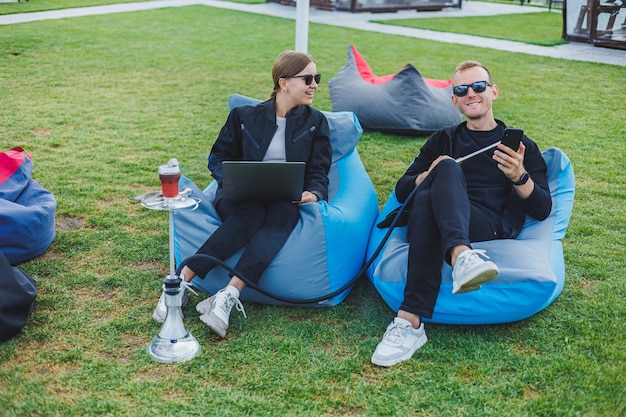 A young woman and a man are sitting in a park on a bean bag and smoking hookah Happy couple relaxing outdoors with hookah