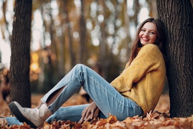 Young woman model sitting in autumn park with yellow foliage maple leaves. Fall season fashion