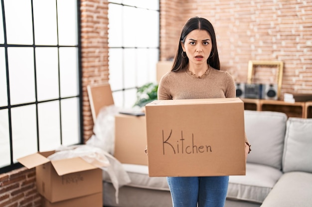 Young woman moving to a new home holding box in shock face looking skeptical and sarcastic surprised with open mouth