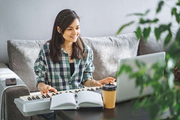 Young woman music teacher playing electric piano teaching remotely using laptop while working from home. Online education and leisure concept.