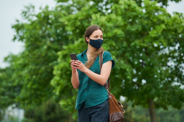 A young woman in a navy blue medical face mask holding a smartphone with both hands while walking in the park