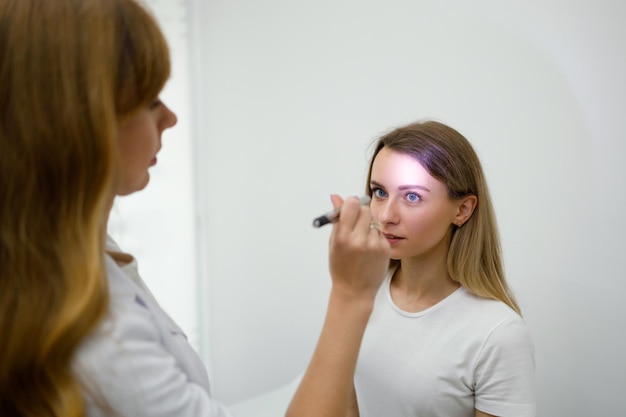 Young woman in neurologist appointment Female doctor checks eyes reflexes with flashlight