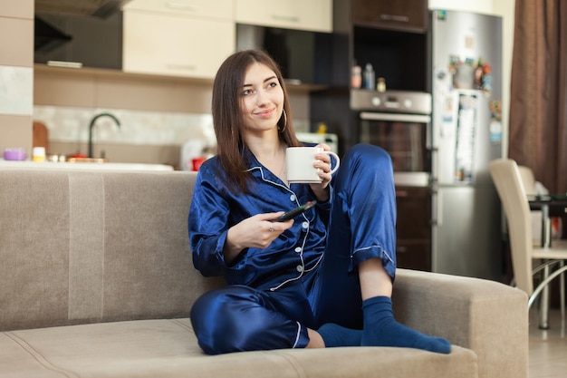 Young woman in nightie drinks coffee and watches TV while sitting on the sofa in the living room