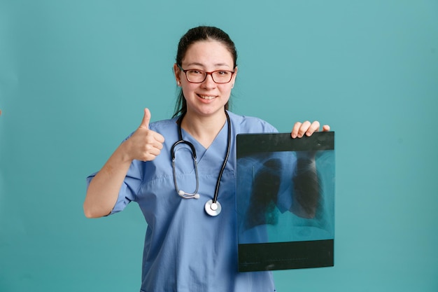 Young woman nurse in medical uniform with stethoscope around neck holding lung xray looking at camera with happy face smiling showing thumb up standing over blue background