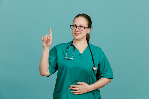 Young woman nurse in medical uniform with stethoscope around neck looking at camera smiling confident showing index finger having good idea standing over blue background