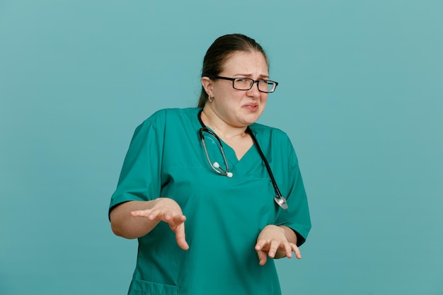 Young woman nurse in medical uniform with stethoscope around neck looking at camera with disgusted expression making defense gesture with hands standing over blue background