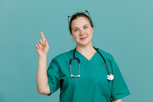 Young woman nurse in medical uniform with stethoscope around neck looking confident smiling showing index finger standing over blue background