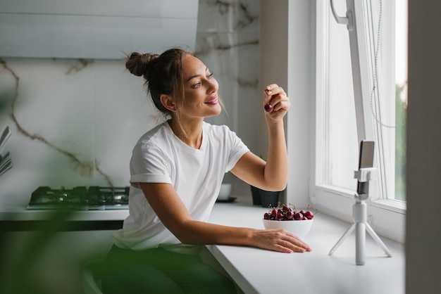 Young woman nutritionist blogger conducts a consultation or an open lesson by video link telling about the benefits of fresh berries sitting in the kitchen with a plate of cherries