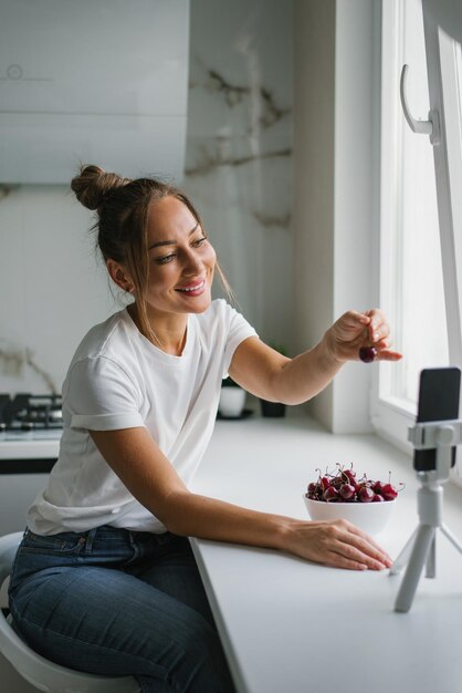 Young woman nutritionist blogger conducts a consultation or an open lesson by video link telling about the benefits of fresh berries sitting in the kitchen with a plate of cherries