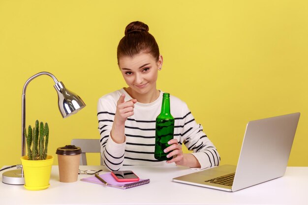 Young woman office employee holding beer bottle in hands looking at pointing to camera with smile