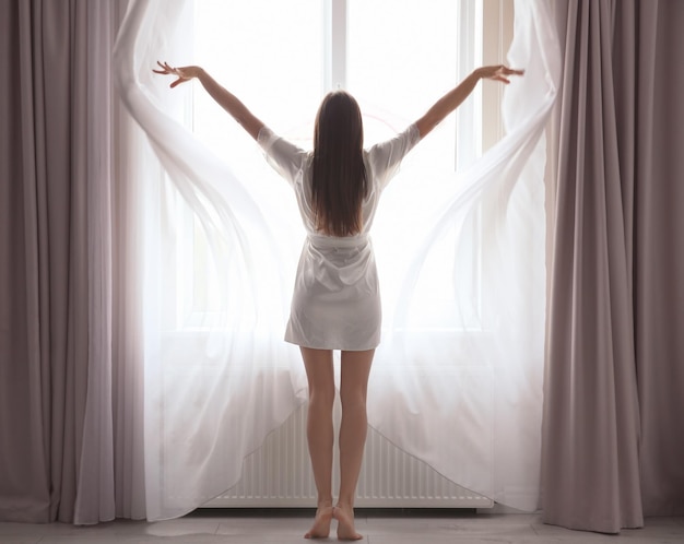 Young woman opening curtains at home