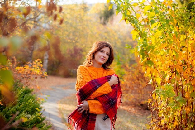 A young woman in an orange sweater wraps herself in a scarf in an autumn park