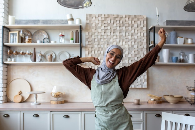 Young woman of oriental origin housewife in hijab in apron cooks and cleans in kitchen she dances