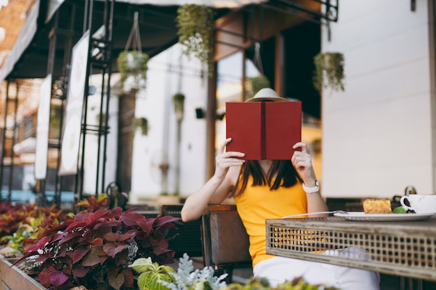 Young woman in outdoors street coffee shop cafe sitting at table in hat, covering face behind red book hiding, relaxing in restaurant during free time