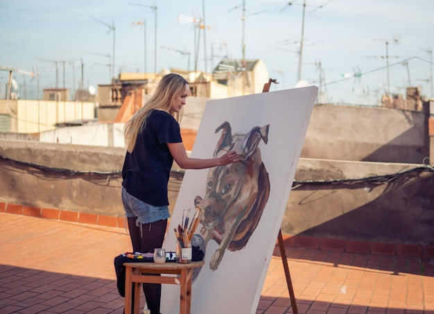 Young woman paint artist drawing at home roof. Picture of bulldog on big canvas. Outdoors art