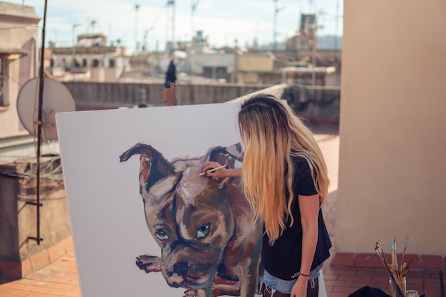 Young woman paint artist drawing at home roof. Picture of bulldog on big canvas. Outdoors art