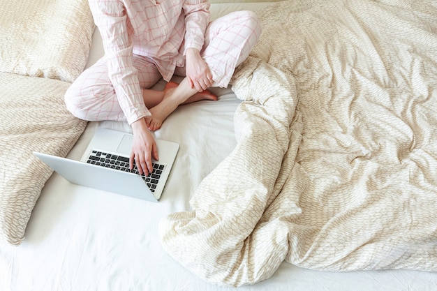 Young woman in pajamas sitting on bed at home working using on laptop pc computer