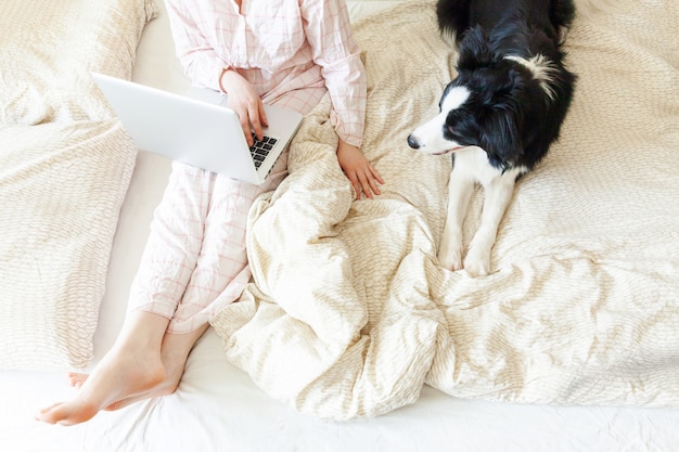 Photo young woman in pajamas sitting on bed with pet dog working using on laptop at home