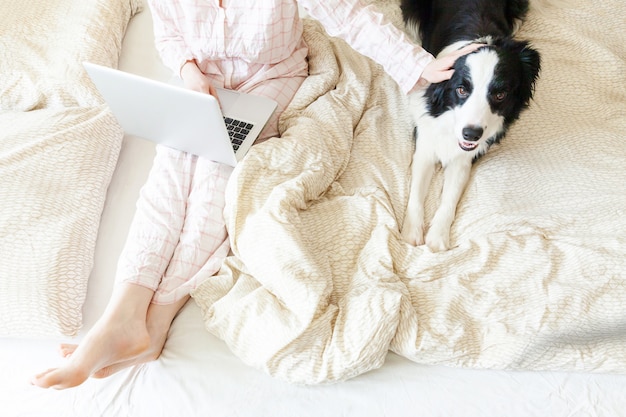 Photo young woman in pajamas sitting on bed with pet dog working using on laptop at home