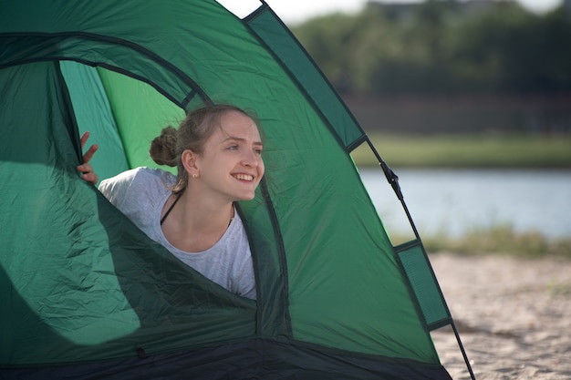 Young woman peeking out of touristic tent and smiling. Morning at campsite.