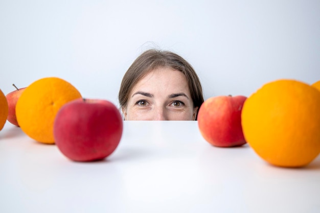 Young woman peeks out from under a table on which fruits lie against a white wall