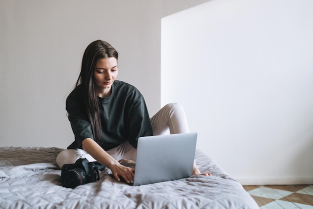 Young woman photographer with digital camera freelancer in casual clothes using laptop sitting on bed at the home