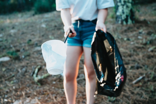 Young woman picking up trash with garbage tongs pointing at cameraForest backgroundPlastic cups and garbage bagNew generationRecycle protecting the planet environmental conservationvolunteer