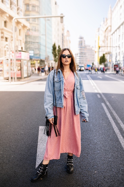 Young woman in an pink dress, denim jacket, boots and eye cat sunglasses standing at the famous view of Gran Via main broadway road in the downtown of Madrid, Spain .