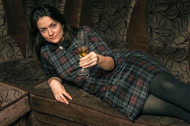 A young woman in a plaid dress drinks wine from a glass lying on a home sofa