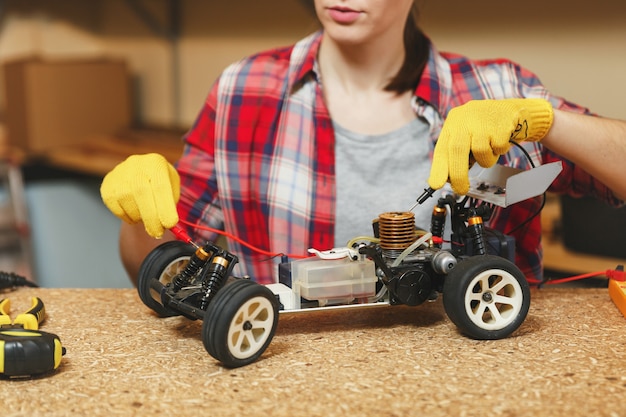 Photo young woman in plaid shirt, gray t-shirt, yellow gloves making toy car iron model constructor, working in workshop at wooden table place with different tools. close up multimeter for electrician.