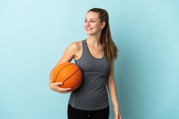 Young woman playing basketball isolated on blue looking to the side and smiling