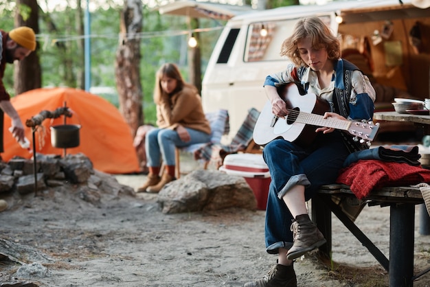 Young woman playing guitar while resting on a camp with her friends in the forest