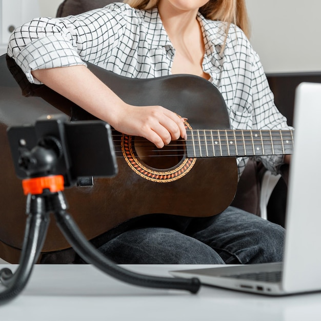 Young woman plays acoustic guitar at home for online audience on laptop and smartphone Online musical guitar performance records via smartphone Female hands closeup