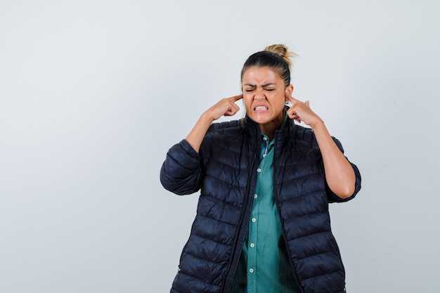 Young woman plugging ears with fingers in shirt, puffer jacket and looking irritated. front view.