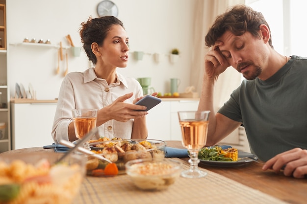 Young woman pointing at mobile phone and talking to man during dinner in the kitchen at home